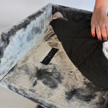 Eine Nahaufnahme eines plüschigen Haustierbettes mit einem weichen, grauen und blauen Stoff. Eine Hand ruht auf einem schwarzen Kissen im Inneren des Bettes.