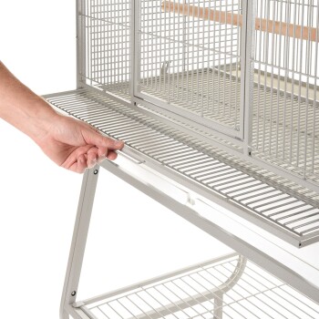 Eine Hand zieht ein herausnehmbares Tablett aus einem großen, grauen Metallvogelkäfig mit mehreren Abteilungen und einer Holzstange im Inneren.