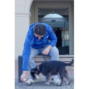 Mann in blauer Jacke sitzt auf einer Bank und bietet einem kleinen schwarz-grauen Hund an der Leine eine Metallschale an; weißes Logo auf der Jacke.