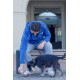 Mann in blauer Jacke sitzt auf einer Bank und bietet einem kleinen schwarz-grauen Hund an der Leine eine Metallschale an; weißes Logo auf der Jacke.