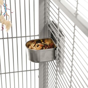 Eine Edelstahlnapf, gefüllt mit gemischtem Haustierfutter, ist an der Seite eines grauen Metallkäfigs mit vertikalen Stäben befestigt.
