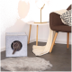 Eine graue Katze greift spielerisch aus einem Filz-Würfel mit einer runden Öffnung, der auf einem gefliesten Boden neben einem gemütlichen Stuhl und einem flauschigen Teppich steht.
