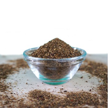 Eine klare Glasschüssel gefüllt mit einem Haufen braunem Kräuter-Haustierergänzungspulver, umgeben von verstreuten Stücken des gleichen Pulvers.