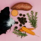 Schwarz-weißer Hund schnüffelt verschüttete runde braune Leckerlis aus einer umgekippten Dose mit der Aufschrift 'DROPS' vor rosa Hintergrund.