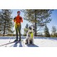 Eine Person in einer orangefarbenen Jacke und grünen Hosen steht im Schnee mit Skistöcken neben einem sitzenden Hund in einem gelben Geschirr, mit Kiefern im Hintergrund.
