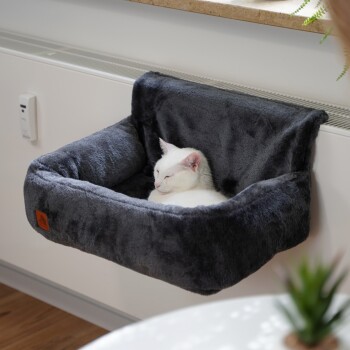 Eine weiße Katze schläft friedlich in einem gemütlichen, dunkelgrauen, wandmontierten Haustierbett, mit einer Pflanze im Vordergrund.