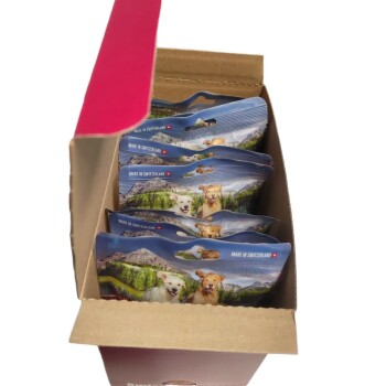 Une boîte en carton contenant plusieurs sachets de nourriture pour animaux de compagnie, avec des images de chiens et des paysages pittoresques, étiquetée "Made in Switzerland."