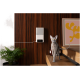 Eine graue Katze mit großen gelben Augen steht auf einer Holztreppe neben einer Topfpflanze, mit einem modernen Futterspender, der an der Wand hinter ihr montiert ist.
