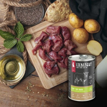 LIEBESGUT Hundefutterdose mit Aufschrift 'Schaf für Hunde' neben rohem gewürfeltem Lamm, Kartoffeln, Öl und Samen auf einem Holzbrett.