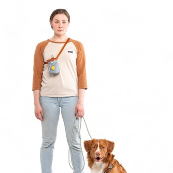 Eine Person in einem beigen und braunen Hemd und hellblauen Jeans steht mit einem goldbraunen Hund an der Leine, der ein blaues Geschirr trägt.