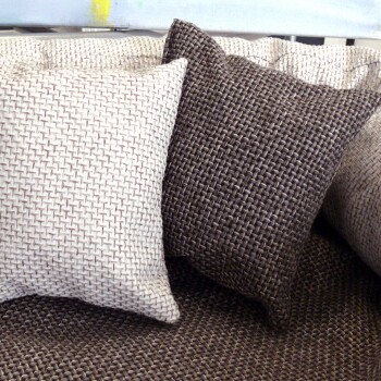 Zwei strukturierte Wurfkissen, eines in Cremefarbe mit einem gewebten Muster und eines in Dunkelbraun, auf einer passenden gewebten Stoffoberfläche ruhend.