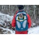 Eine Person in einer roten Jacke trägt eine Katze in einem blauen Kurgo-Rucksack, während die Katze einen grünen Pullover trägt, umgeben von schneebedeckten Bäumen.