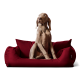 Ein hellbrauner Hund sitzt aufrecht auf einem plüschigen roten Haustierbett mit zwei dekorativen Kissen und schaut neugierig zur Seite.