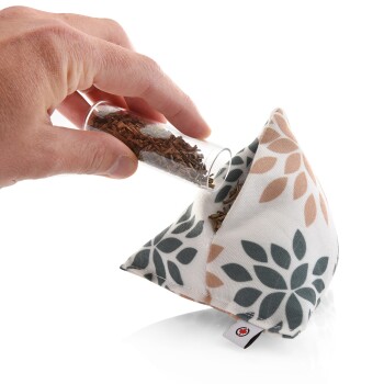 Eine Hand, die braune Kräuter aus einem kleinen Glasrohr in einen dekorativen Stoffbeutel mit floralem Muster in Beige und Grün gießt.