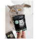 Eine graue Katze mit auffälligen orangen Augen schaut in die Kamera, während eine Hand einen Beutel mit "CAT'S LOVE" Katzenfutter hält, auf dem eine weiße Katze abgebildet ist.
