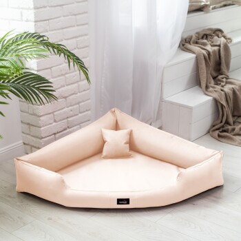 Ein dreieckiges Haustierbett aus weichem Pfirsichstoff, mit einem kleinen passenden Kissen, platziert in einem hellen Raum mit einer Pflanze und einer gemütlichen Decke.