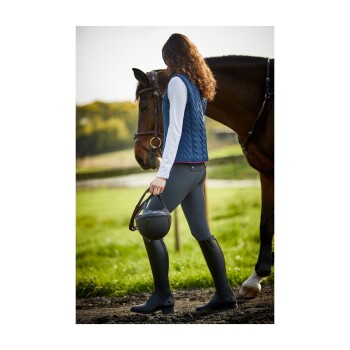 Eine Frau in einer marineblauen gesteppte Weste und Reitstiefeln geht neben einem braunen Pferd auf einer grasbewachsenen Fläche, während sie einen Reithelm hält.