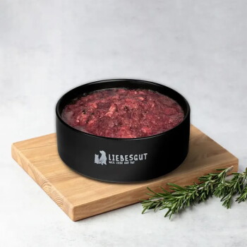 Schwarzer Napf mit der Aufschrift 'LIEBESGUT - WEIL LIEBE GUT TUT', gefüllt mit rohem Hackfleisch, auf einem Holzbrett mit Rosmarinzweig