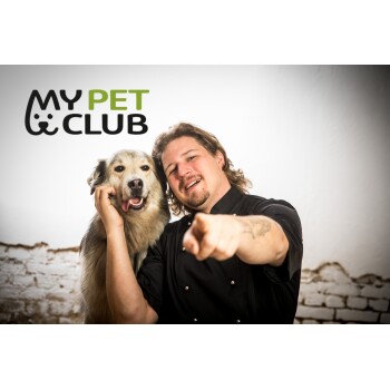 Eine Person in einer schwarzen Kochjacke zeigt auf die Kamera, während sie einen kleinen Hund hält. Das Logo "MY PET CLUB" wird deutlich darüber angezeigt.