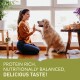 A woman kneels on a wooden floor with a golden retriever in a bright room. Text overlay: "PROTEIN RICH, NUTRITIONALLY BALANCED, DELICIOUS!"