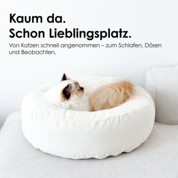 Zwei Katzen zusammengerollt in einem weißen runden Katzenbett auf einem Sofa; sichtbarer Text: "Kaum da. Schon Lieblingsplatz."