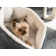 Ein kleiner Hund, der sich in einem gemütlichen, plüschigen Hundebett mit grauem Äußeren und weichem weißen Innenraum zusammengekuschelt hat und neugierig in die Kamera schaut.