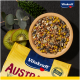 Eine Holzschüssel mit bunten Samen und Körnern auf einer dunklen Oberfläche, mit Kiwi-Scheiben, grünen Blättern und einer gelben "Vitakraft AUSTRALIA" Tüte.