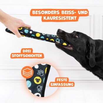 Ein schwarzer Hund zieht verspielt an einem bunten Kauspielzeug, das von einer Person gehalten wird, mit Text über Haltbarkeit und Materialien.