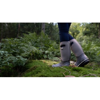 Nahaufnahme der Beine einer Person, die stilvolle graue Gummistiefel trägt, auf moosigem Boden stehend, umgeben von üppigen grünen Farnen in einem Wald.