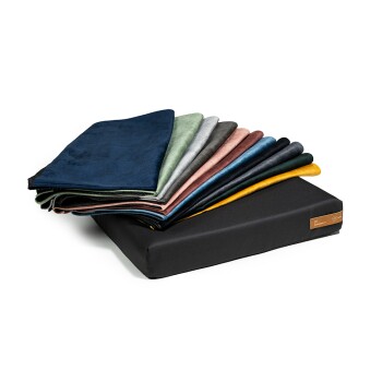 Ein schwarzes Haustierbett mit einem Stapel bunter, weicher Decken in verschiedenen Farbtönen, darunter Blau, Grün, Grau und Gold, oben drauf.