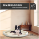 Ein schwarz-weißer Hund entspannt sich auf einem gemütlichen Bett in einem hellen Raum mit Holzböden und einem Tisch. Text: "ZUM EINKUSCHELN schafft ein gemütliches Ambiente."