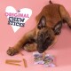 Een speelse puppy die op een kauwstaafje bijt, omringd door verschillende kauwstaafjes en twee verpakkingen gelabeld 'Original Chew Sticks' op een roze achtergrond.