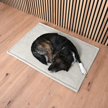 Ein eingerollter Hund ruht auf einer weichen, hellen Haustiermatte auf einem Holzboden, mit einer modernen Holzlattenwand im Hintergrund.