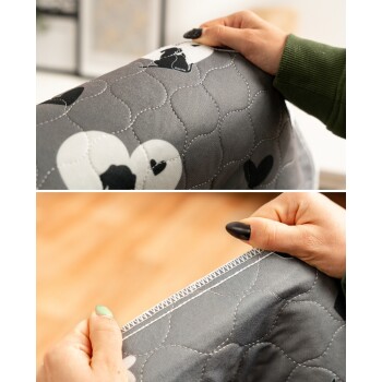 Nahaufnahme von Händen, die eine graue gesteppte Haustierdecke mit schwarzen Katzen- und Herzmustern sowie sichtbaren Nahtdetails halten.