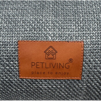 Braunes Lederetikett mit Pfoten-im-Haus-Logo und der Aufschrift 'PETLIVING' sowie 'place to enjoy' auf grauem gewebtem Stoff.