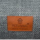 Braunes Lederetikett mit Pfoten-im-Haus-Logo und der Aufschrift 'PETLIVING' sowie 'place to enjoy' auf grauem gewebtem Stoff.