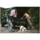 Ein weißer Hund mit einer orangefarbenen Weste sitzt auf einer Matte neben einem schwarzen SUV mit offenem Kofferraum. Zwei Personen bereiten Angelausrüstung vor.