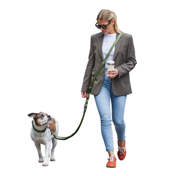 Frau in Blazer und Jeans geht mit einem braun-weißen Hund an einer grünen Leine spazieren und hält einen Reise-Kaffeebecher.