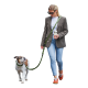 Frau in Blazer und Jeans geht mit einem braun-weißen Hund an einer grünen Leine spazieren und hält einen Reise-Kaffeebecher.