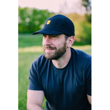 Person mit dunkelblauer Baseballkappe mit hellbraunem Patch und dunkelblauem T‑Shirt sitzt auf einer Wiese; Gesicht verdeckt