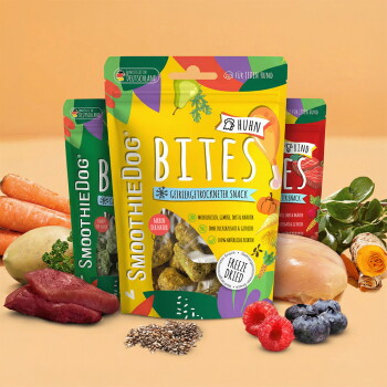 Drei bunte SmoothieDog BITES gefriergetrocknete Snack-Beutel (Huhn), umgeben von Fleisch, Früchten, Gemüse und Samen.