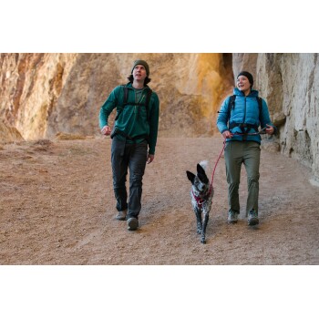 Zwei Männer, die auf einem Schotterweg in einem felsigen Gebiet gehen, begleitet von einem schwarz-weißen Hund an der Leine.