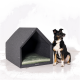 Ein schwarz-brauner Hund sitzt neben einem modernen, grauen Filz-Hundehäuschen mit einem grünen Kissen darin, vor einem weißen Hintergrund.