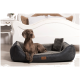 Ein brauner Hund ruht auf einem plüschigen, dunkelgrauen Hundebett mit einem kleinen Kissen, in einem gemütlichen Raum mit einem weißen Bett und floraler Dekoration.