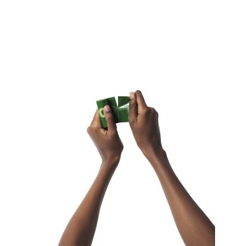 Une paire de mains tenant un paquet de produit pour animaux de compagnie vert, partiellement ouvert, sur un fond blanc.