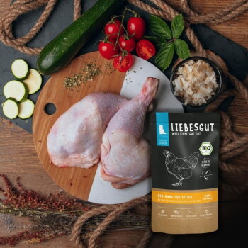Tierfutterbeutel "LIEBESGUT" (Bio-Huhn für Kitten) neben rohen Hähnchenschenkeln auf einem Schneidebrett mit Tomaten, Zucchini und Kräutern.