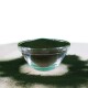 Eine Glasschüssel gefüllt mit grünem Spirulina-Pulver, umgeben von einer Staubschicht des gleichen Pulvers auf einer weißen Oberfläche.