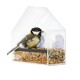 A small bird, possibly a great tit, perched on a black bar inside a transparent bird feeder filled with various seeds.