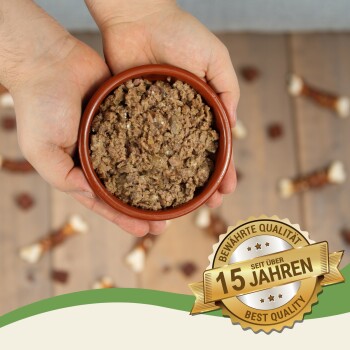 Eine Person hält eine kleine braune Schüssel mit Hundefutter, umgeben von Hundeleckerlis, mit einem Abzeichen, das "15 Jahre Beste Qualität" liest.