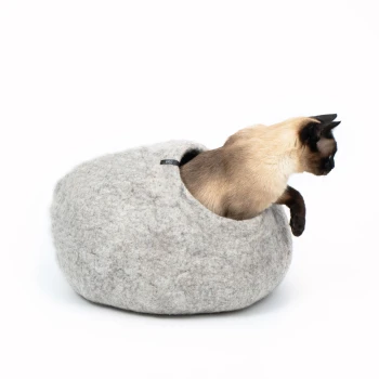 Eine Siamkatze schaut aus einem gemütlichen, grauen Filz-Hundebett mit runder Form und kleiner Öffnung heraus.